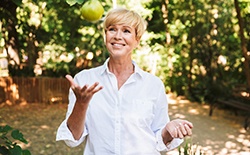 An older woman wearing a white shirt and tossing an apple, happy with her new, functional smile