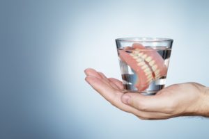 hand holding a glass of water with two full dentures soaking in it 
